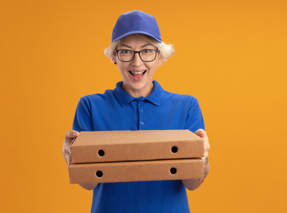 微笑身着蓝色制服 戴着眼镜 拿着比萨饼盒的快乐的年轻送货员在橙色的墙上欢快地微笑着欢呼拿着送货