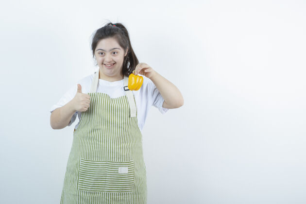 食物穿着格子围裙的年轻漂亮女孩 手里拿着黄色的甜椒 竖起大拇指残疾成人年轻人