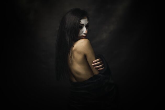 脸半裸的女人脸上涂着小丑妆站在黑色背景前美女幽默服装