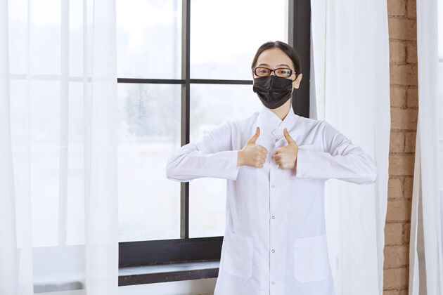 女性戴着黑面具和眼镜的医生站在窗边 展示着享受的标志细菌喜欢病毒