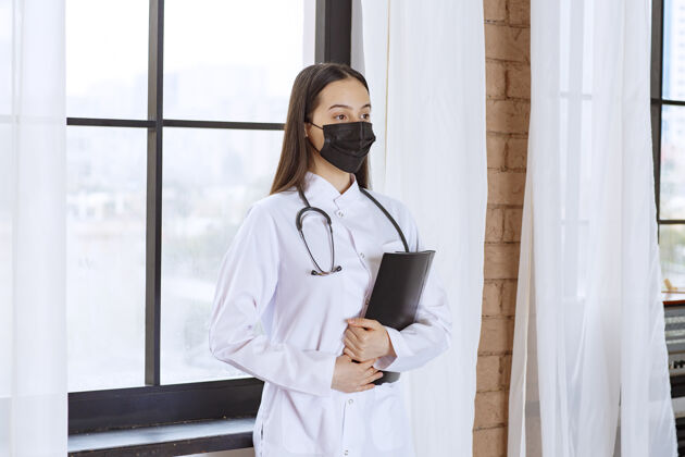 医疗医生拿着听诊器和黑色口罩站在窗口旁边 手里拿着一个黑色的病人病史文件夹感染诊所病毒