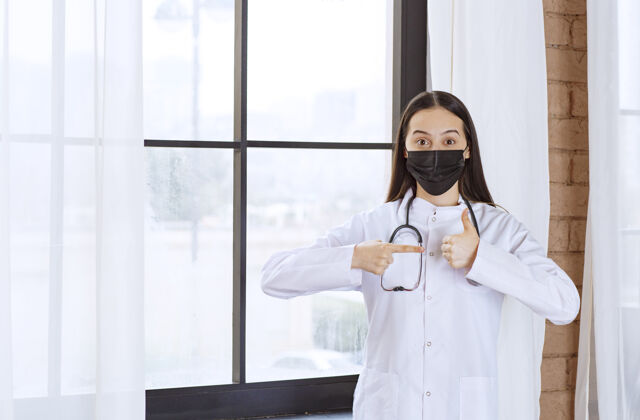 诊所戴着黑色口罩的医生站在窗边 带着听诊器 并显示出满意的迹象细菌病毒医学
