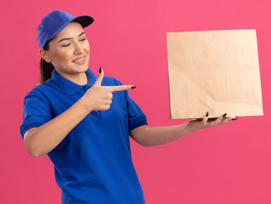帽子身穿蓝色制服 戴着帽子的年轻送货员拿着纸包 食指指着纸包 自信地微笑着站在粉色的墙上纸手指指着