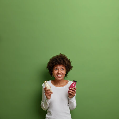 非洲正卷曲的女人拿着冰沙和素食牛奶 喝排毒饮料 有着均衡的适当营养 集中在上面带着灿烂的笑容 看到一些令人愉快的东西 隔离在绿色的墙上青少年室内水果