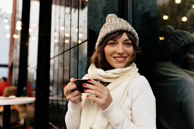 商业女性美丽可爱的女人 带着灿烂的笑容 戴着针织帽 穿着毛衣 端着一杯咖啡 在外面享受着咖啡时光高质量的照片大声工作学习