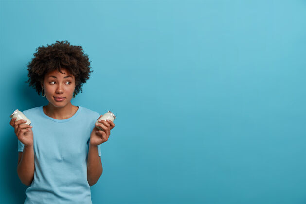 年轻健康的运动型黑人头发女人 拿着一瓶新鲜酸奶 准备早餐 营养充足 注意力集中 穿着休闲服 对着蓝色的墙壁摆姿势肖像乳制品民族