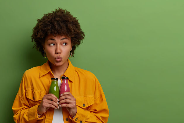 年轻人体贴美丽的美国黑人妇女手持玻璃瓶新鲜的苹果和草莓冰沙 引领健康的生活方式 喝营养有机饮料来保持健康 隔离在绿色的墙上肖像饮食奶昔