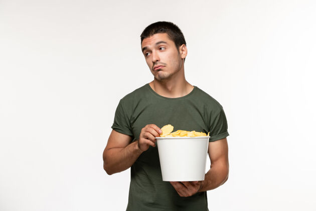 灯光正面图身穿绿色t恤的年轻男性 带着土豆cips 在浅白的墙上看电影 人男孤独的电影院观看土豆容器
