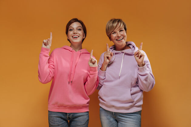 女儿有趣的两位女士 短而酷的发型 穿着现代的粉色连帽衫和牛仔裤 在橙色孤立的背景下竖起大拇指肖像老年人连帽衫