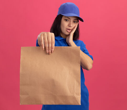 制服穿着蓝色制服 戴着帽子 拿着纸包裹的年轻送货女孩站在粉红色的墙上看着它 惊讶和惊讶交货举行包装