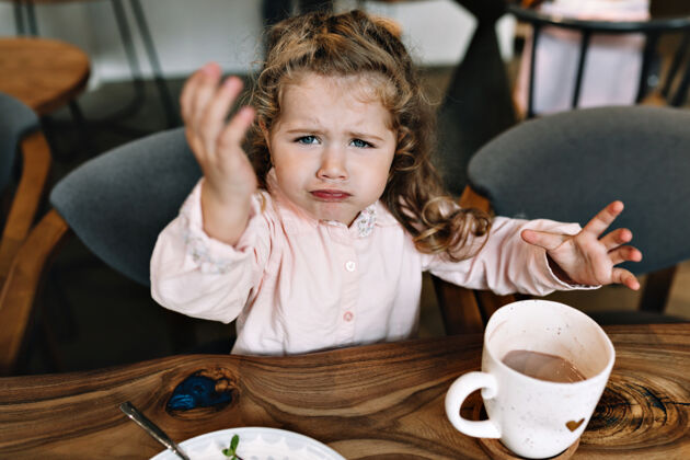 漂亮悲伤的小女孩正坐在餐馆的桌子旁抱着坐着早餐