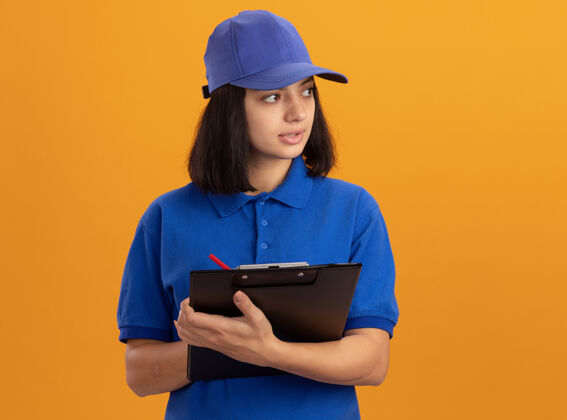 年轻身穿蓝色制服 戴着帽子的年轻送货员拿着空白页和铅笔的剪贴板 严肃的脸站在橙色的墙上 看着一旁靠边递送页面