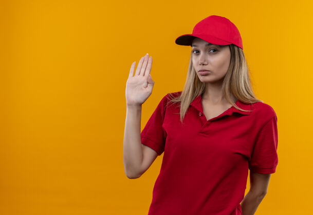 年轻看着镜头 穿着红色制服 戴着帽子的年轻送货女孩在橙色背景下显示出停止的手势穿相机制服