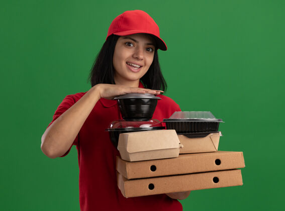 微笑身着红色制服 头戴鸭舌帽 手持披萨盒和食品包的快乐年轻送货女孩站在绿色的墙上 开心地微笑着送货女孩食物