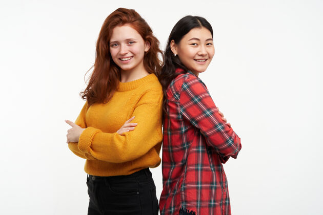 毛衣两个年轻的女性朋友穿着黄色毛衣和格子衬衫背靠背站着 双臂交叉人和生活方式的概念隔着白墙孤立两个衬衫介绍