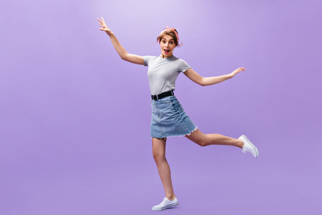 年轻穿着裙子和衬衫的漂亮女人在紫色的背景上跳跃穿着白色运动鞋的时髦发型的有趣的年轻女孩摆姿势休闲一件肖像