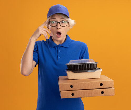 盒子身穿蓝色制服 戴着眼镜 拿着比萨饼盒和食品包的年轻送货员惊讶地越过橙色的墙壁送货帽子年轻人