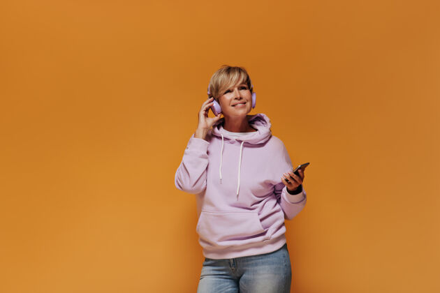 橙色背景一位面带微笑的时髦老太太 金发碧眼 穿着粉色运动衫和浅牛仔裤 戴着淡紫色耳机和智能手机成熟中电话