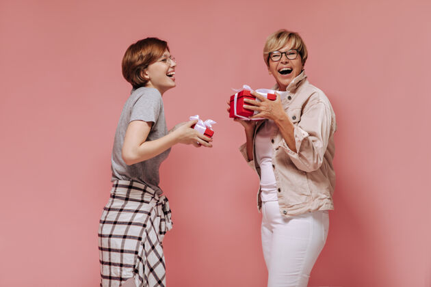 成熟快乐的两位短发女士 戴着酷酷的眼镜 穿着时髦的衣服 笑着拿着粉色背景的红色礼盒灰色短发休闲