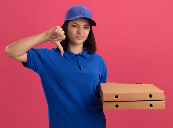 展示身穿蓝色制服 头戴鸭舌帽 拿着比萨饼盒的年轻送货员不高兴地竖起大拇指站在粉红色的墙上送货披萨拿着