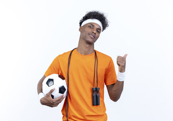 壁板自信的年轻美国黑人运动型男子戴着头带和腕带 肩上拿着跳绳的球 在白色背景上孤立地指向侧面 留有复制空间肩膀非裔美国人运动