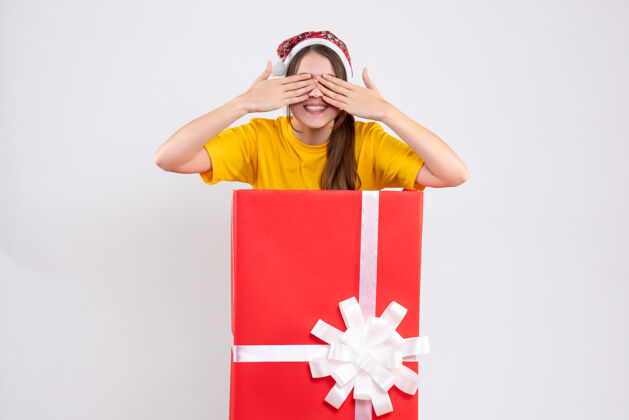大正面图可爱的女孩戴着圣诞帽遮住眼睛 双手站在圣诞礼物后面证人圣诞老人购物