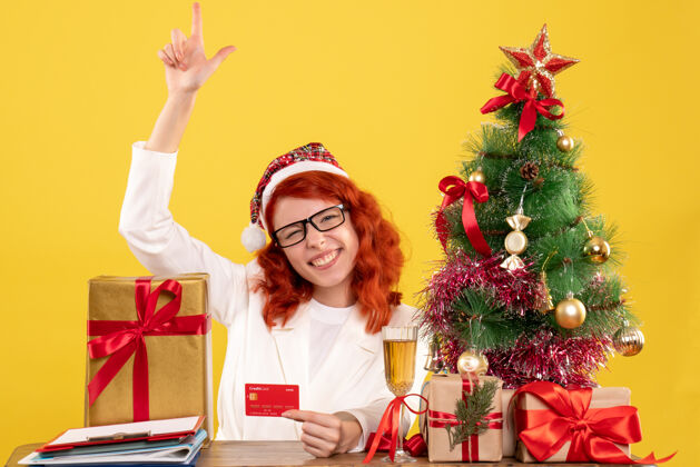 圣诞节前视图女医生拿着银行卡在圣诞礼物和圣诞树周围庆祝礼物年份