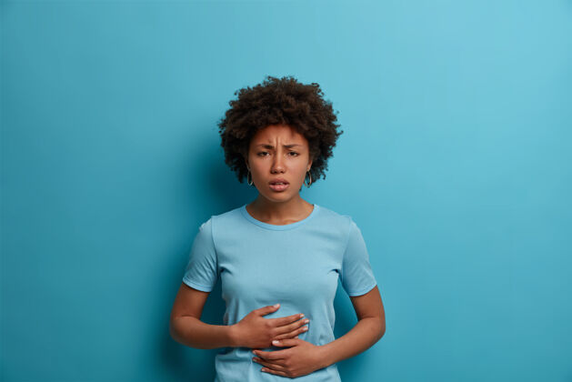 不适不高兴的非洲女人感到胃部不适 手掌放在肚子上 经期抽筋 穿休闲t恤 吃变质的食物 孤立在蓝色的墙上 用模仿来表达负面情绪年轻疾病非洲