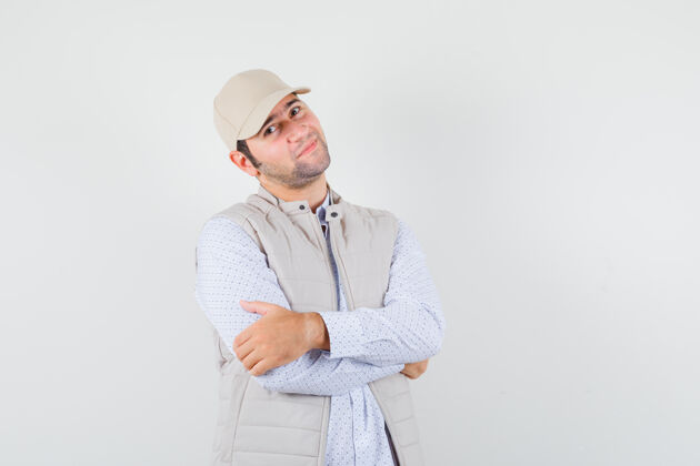 男人年轻人穿着衬衫 无袖夹克 戴着帽子 双臂交叉着站着 看上去很满足 前视图优雅年轻帽子
