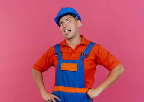 穿看着摄像机 穿着制服 戴着安全帽的年轻男性建筑工人露出舌头 双手放在臀部 粉色背景上孤立着手制服相机