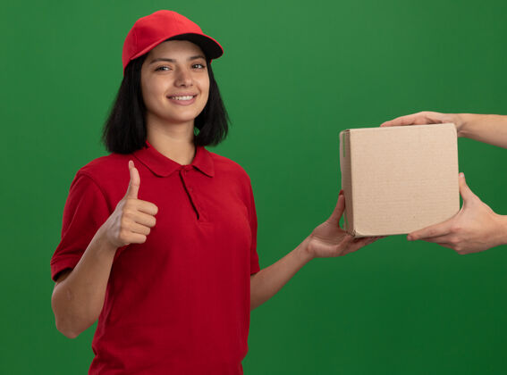 拇指身着红色制服 头戴鸭舌帽的年轻送货员站在绿色的墙上 友好地微笑着向顾客竖起大拇指顾客制服帽子