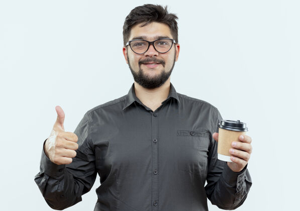 戴着高兴的年轻商人戴着眼镜拿着一杯咖啡 他的大拇指孤立地竖在白色背景上拿着年轻人请