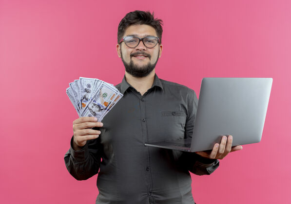 笔记本电脑高兴的年轻商人戴着眼镜拿着笔记本电脑和现金隔离在粉红色的背景上现金请商务人士