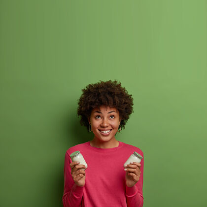 乳制品卷曲正面女人的垂直镜头微笑着俯视 手持美味的白色有机酸奶 穿着休闲的粉色套头衫 对着绿色的墙壁摆姿势 向上腾出空间健康营养自制肖像请