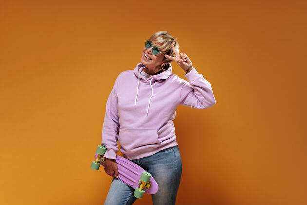 女士迷人的酷女人 金色的现代发型 戴着绿色眼镜 粉色超大连帽衫 微笑着 展示着和平的标志 在橙色背景下摆出滑板的姿势年龄老年人连帽衫