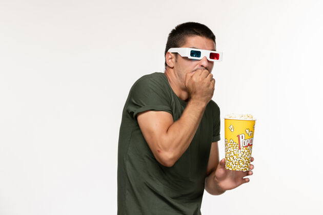 爆米花正面图身穿绿色t恤的年轻男子戴着d型太阳镜拿着爆米花在白墙上看电影孤独电影院男电影电影电影观看