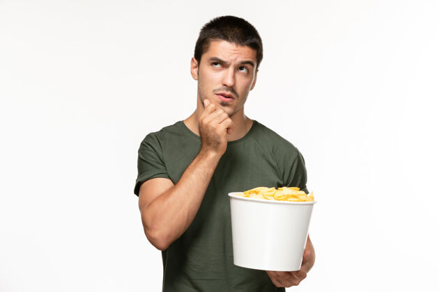 观点正面图身穿绿色t恤的年轻男子手持土豆片 在白墙上看电影思考孤独的电影成人电影男人