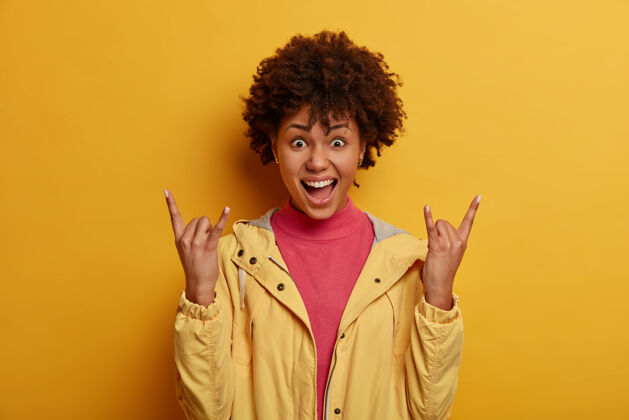 休闲积极的卷发女人在很棒的音乐会上 享受派对 做摇滚手势 重金属标志 摇滚歌迷 穿着夹克 隔离在黄色的墙上 感觉无忧无虑表情氛围非洲