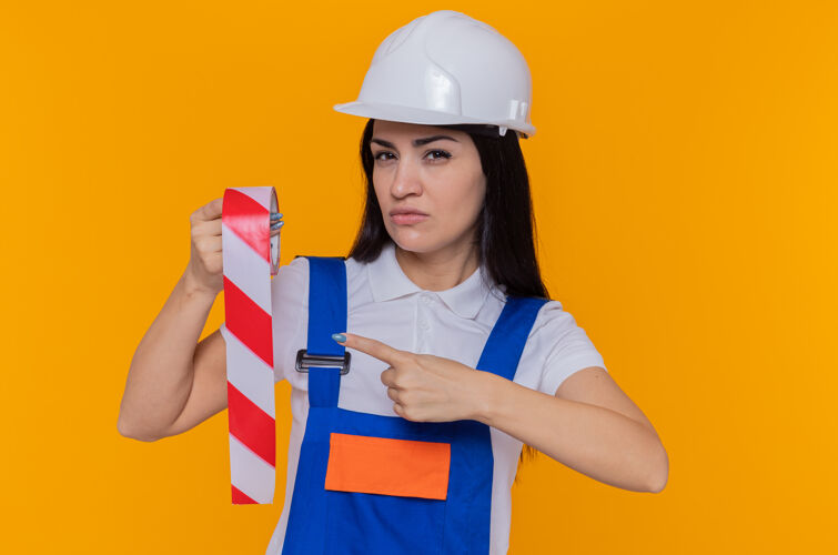 手指年轻的建筑工人 穿着建筑制服 戴着安全帽 拿着胶带 用食指指着它 严肃的脸站在橙色的墙上看着前面头盔女人站着