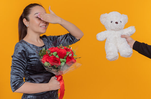 欢呼快乐的亚洲女人看着快乐和惊讶蒙着眼睛接受泰迪熊作为礼物微笑着欢庆国际妇女节站在橙色的墙上手掩护快乐