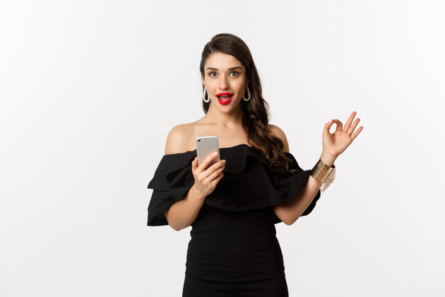 服装网上购物概念穿着时髦的黑色连衣裙 化妆 显示同意登录并使用手机应用程序 白色背景的女人美丽电话女人