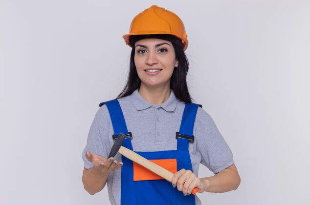 女人身穿建筑制服 头戴安全帽的年轻建筑女工人挥舞着锤子 面带微笑自信地站在白色的墙上看着前方建筑自信年轻