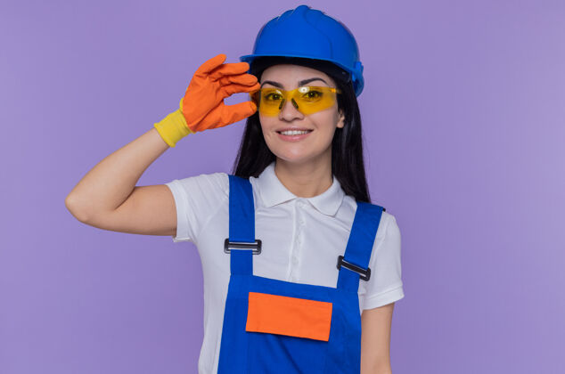 快乐快乐的年轻建筑妇女穿着建筑制服 戴着安全帽 戴着橡胶手套 面带微笑 自信地站在紫色的墙上自信手套建筑工人