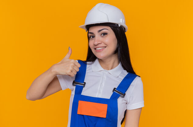拇指身穿施工制服 头戴安全帽的快乐的年轻建筑女工人站在橙色的墙上 面带微笑地竖起大拇指看着前方建筑站立欢呼