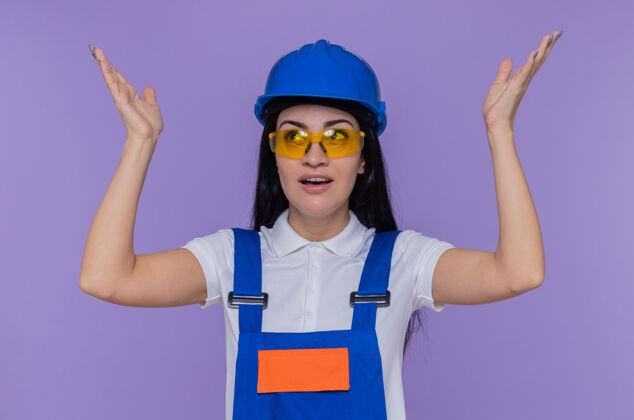 手臂身穿施工制服 头戴安全帽的年轻建筑女工人抬起双臂站在紫色的墙壁上 既高兴又惊讶安全惊喜女人