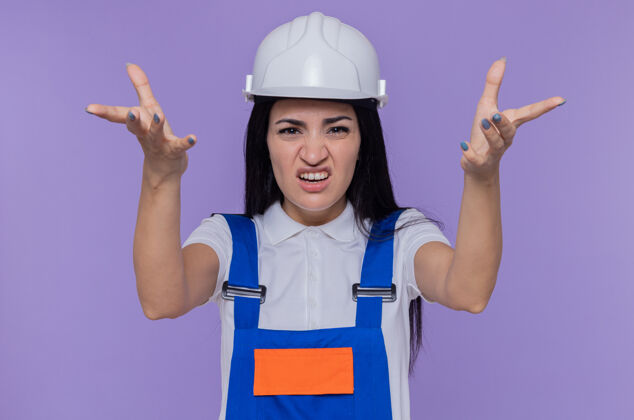制服身穿施工制服 头戴安全帽的年轻建筑工人站在紫色的墙上 双臂高举 像是在争吵建筑工人站立头盔