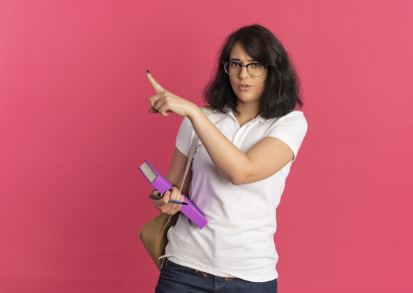 背部年轻恼怒的漂亮白人女学生戴着眼镜 背着书包 拿着笔和书指着粉色的侧面和复印空间穿复制包