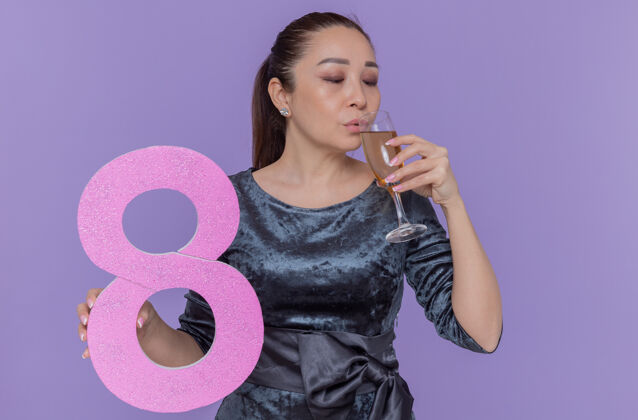快乐快乐的亚洲女人拿着八号硬纸板和香槟酒杯 站在紫色的墙上庆祝国际妇女节八喝酒制造