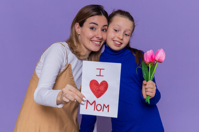 花快乐的女儿站在紫色的墙上 给她惊喜而微笑的母亲送上贺卡和郁金香花 庆祝母亲节庆祝惊喜女儿