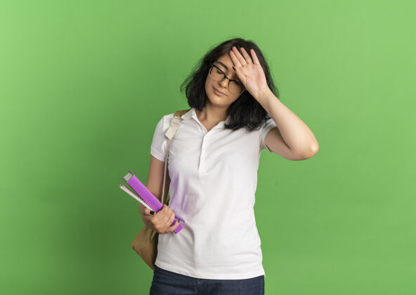 复制年轻疲惫漂亮的白人女学生戴着眼镜 背着书包 手放在额头上 拿着书放在绿色的纸上 留着复印空间背空间戴
